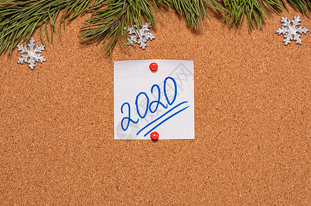 带有 2020 年手写体的白色便签贴在布告板上 布告板上装饰着松树枝和雪花 2020 年圣诞节 新年庆祝季节的概念 特写镜头背景图片