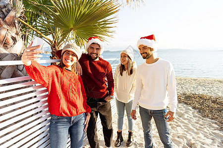 一群戴着圣诞老人帽子的年轻白人朋友在冬天的圣诞节假期在海滩上自拍很开心 — 两对人使用技术分享他们的旅程图片