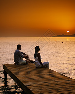 希腊克里特岛 年轻浪漫的情侣在日出时分坐在沙滩上的木码头上拥抱着金色的天空 假期和旅行的概念 浪漫的年轻情侣在海边约会 太阳 夫图片