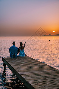 希腊克里特岛 年轻浪漫的情侣在日出时分坐在沙滩上的木码头上拥抱着金色的天空 假期和旅行的概念 浪漫的年轻情侣在海边约会 爱 水图片