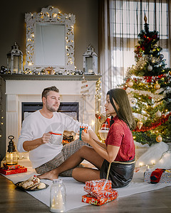可爱的年轻夫妇在壁炉旁放着一棵圣诞树 一家人坐在地板上 在圣诞树附近的夫妇 假期 圣诞彩灯图片