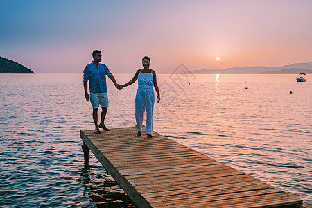 希腊克里特岛 年轻浪漫的情侣在日出时分坐在沙滩上的木码头上拥抱着金色的天空 假期和旅行的概念 浪漫的年轻情侣在海边约会 水 坐着图片