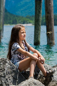 一位可爱的小姑娘坐在阿尔卑斯湖的岸边 在奥地利Plan See湖 帽 周末图片