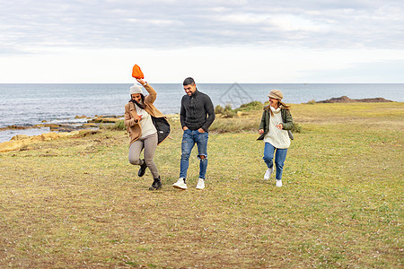 三个混血无忧无虑的朋友在户外跑步互相开玩笑 — 千禧一代在海边的草地上玩得开心 在寒假里微笑 穿着衣服在海洋度假胜地享受大自然图片