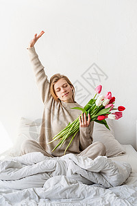 坐在床上的快乐女人穿着睡衣伸着手拿着郁金香花束图片