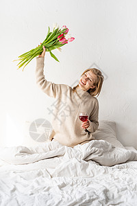 坐在床上的快乐女人穿着睡衣 拿着酒杯和郁金香花花束 葡萄酒 浪漫的图片