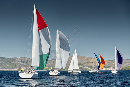 帆船在日落时参加帆船赛 帆船比赛 水面上的帆反射 五颜六色的大三角帆 船尾的船数 岛屿在背景中 天气晴朗 地中海 海浪背景图片
