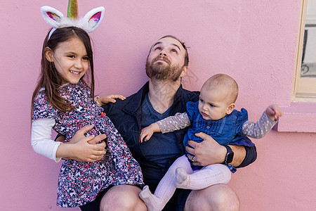 快乐的父亲拥抱他两个可爱的女儿 在粉红色的墙壁前 乐趣 童年图片