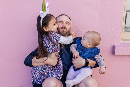 快乐的父亲抱着两个可爱的女儿 带着粉红色的墙做背景 独角兽角 姐姐图片