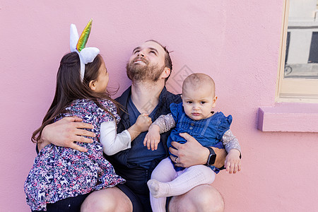 快乐的父亲拥抱他两个可爱的女儿 在粉红色的墙壁前 家庭 男人图片
