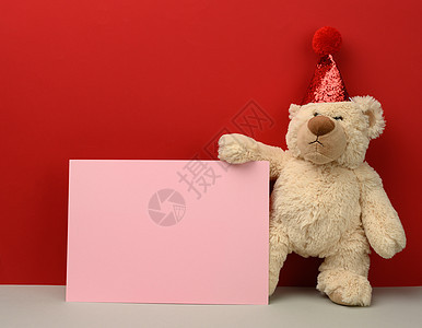 穿着红色喜剧帽的泰迪熊 在红色背景上持有一张粉红纸图片