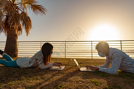两名学生在日落时躺在草地上使用笔记本电脑在热带海洋度假胜地的城市公园学习课程 年轻的自主创业夫妇通过移动技术为自己工作背景图片