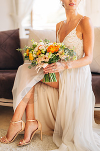 新娘穿着婚礼服和高头凉鞋 坐在房间里的沙发上 手里握着结婚花束图片