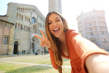 年轻的女游客在意大利帕尔马市笑着自拍 女旅行者在 2020 年意大利文化之都帕尔马的大教堂广场自拍并展示 V 形手势图片