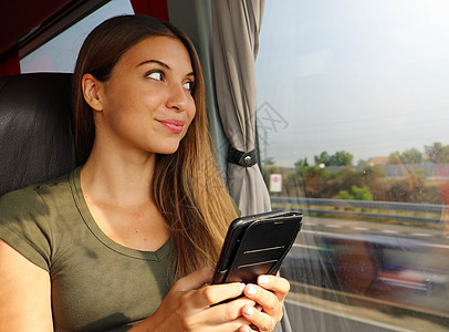 透过公交车窗看年轻漂亮的女人 快乐的公共汽车乘客坐在座位上旅行 手里拿着智能手机透过窗户看图片