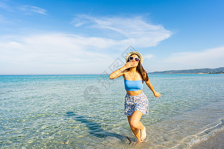 年轻美丽的女士用手亲吻手对嘴 看着在海水中穿戴白帽子和墨镜的照相机走在水里 花时间旅行 争取幸福更好的生活体验 笑声 图片