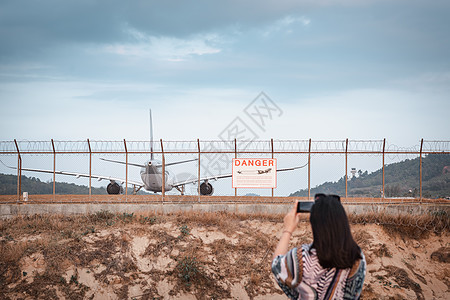 旅游妇女正在拍摄飞机的照片 同时从机场外起飞降落在跑道轨道上 亚洲旅游妇女在机场围栏旁玩摄影车飞机图片