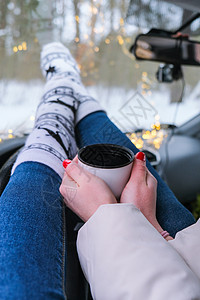 穿着袜子和袜子的女子腿手握着热水杯咖啡或冬车茶 圣诞袜 加兰灯光不亮 脚 司机图片