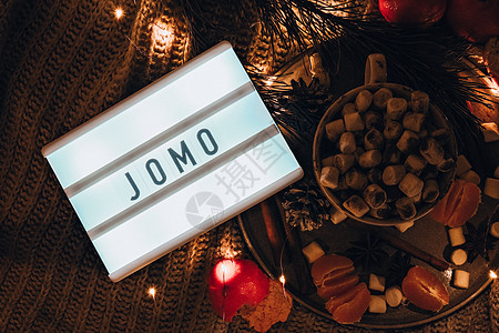 带有文字 JOMO Joy 的灯箱 错过了杯子 床上放着热冬可可和棉花糖橘子云杉树枝 圣诞彩灯 松果装饰 舒适的家 乔莫图片