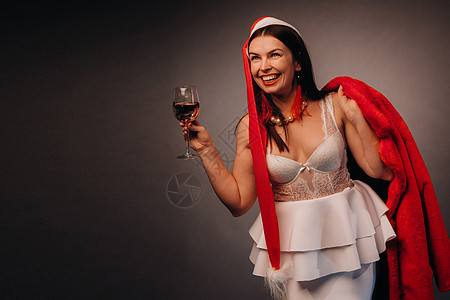 一个戴着圣诞帽和白色裙子的女人 手里拿着玻璃杯和一件红色皮大衣 背景是黑色 圣诞节 假期 深色背景 喝图片