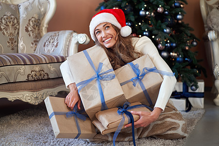 穿着白色毛衣的妇女 圣诞礼帽和牛仔裤 坐在圣诞节树旁边的地板上 带现装箱 圣诞帽 漂亮的图片