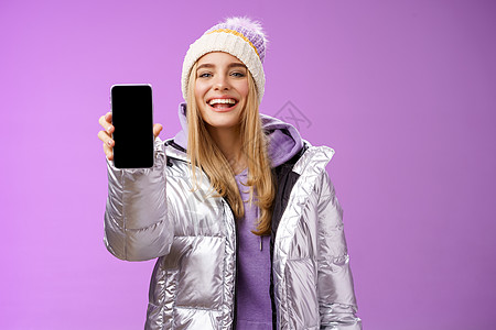 活泼开朗的金发女性推荐使用应用编辑完美的照片 手持智能手机展示手机显示屏 自豪地令人满意的微笑相机 站在紫色背景 互联网 山脉图片