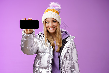 自信友善的金发女孩 身穿户外银色闪闪发光的夹克帽 手持智能手机水平展示手机显示自信的微笑 推荐使用应用程序 紫色背景 情绪 寒冷图片