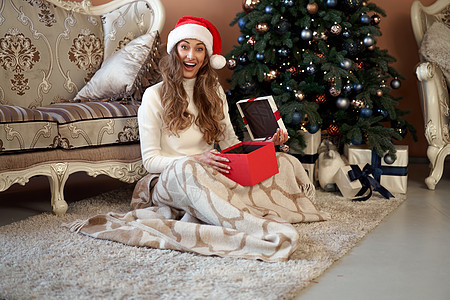 穿着白色毛衣的妇女 圣诞礼帽和牛仔裤 坐在圣诞节树旁边的地板上 带现装箱 惊喜 格子图片