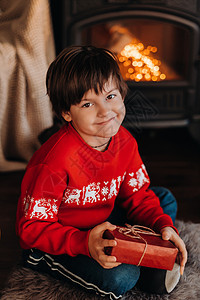 一个笑着男孩的肖像 手拿着礼物 在家壁炉附近 圣诞节图片
