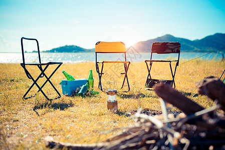 三把火在草地上的三把野餐椅 在前景和湖的营火有山的在背景中 露营和旅行的概念 远足装备主题背景