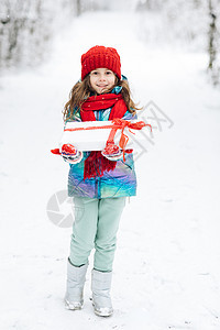 戴着红色帽子的孩子带着圣诞礼物和雪中的礼物 冬季户外乐趣 圣诞节前夕 孩子们在白雪皑皑的公园玩耍 快乐的小白人女孩微笑着拿着礼品图片