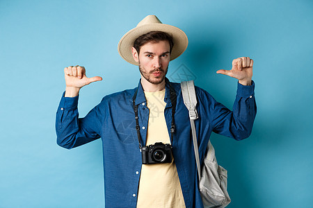 自信的帅哥戴着夏帽 用吹牛的眼神指着自己 去度假旅行 拿着背包和相机 蓝色背景 成熟 大胡子图片