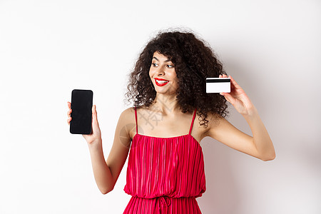 在线购物概念 身着红色礼服 展示塑料信用卡和空移动电话屏幕 站在白背景上高雅的卷卷发女色女子 钱 化妆品背景图片