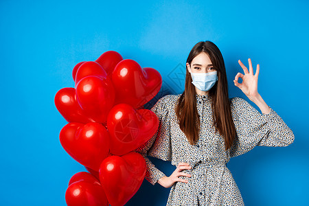 Covid-19 情人节和大流行病概念 美丽的年轻女子面对面罩和着装 在认可时显示OK标志 站在浪漫的红心气球旁边 蓝背景 蓝色图片