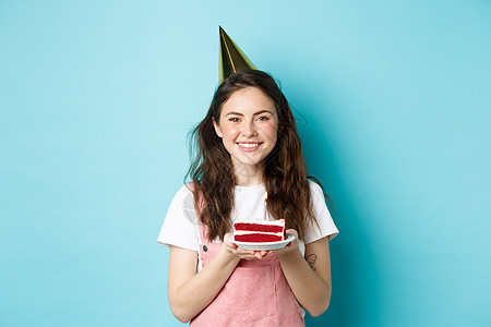 节日和庆祝活动 戴着派对帽的快乐生日女孩拿着生日蛋糕 微笑着 在点燃的蜡烛上许愿 站在蓝色背景下 喜悦 迷人的图片