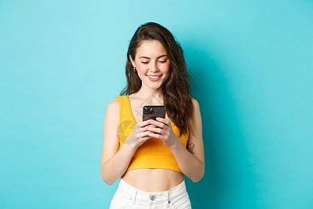 迷人的现代女性在手机上聊天 带着温柔的微笑看着屏幕 在约会应用程序上发消息 站在蓝色背景上图片