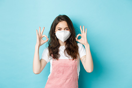 Covid 健康和流行病的概念 满意的戴呼吸器的漂亮女孩 医用面具在批准时显示好的标志 使用预防冠状病毒的措施 蓝色背景图片