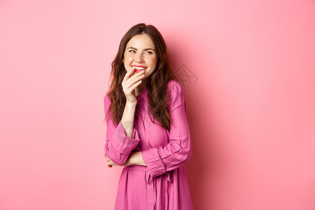 科克特蒂希的年轻女子笑笑 用手指握着嘴唇和小毛孔可爱的脸脸 一边看宣传文本 站在粉红背景上站立 工作室 广告图片