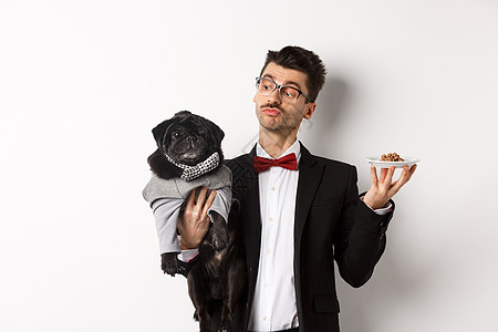 穿着漂亮西装的帅气年轻狗主 拿着可爱的黑瓜和动物食物盘子 站在白色背景上 快乐 生日背景图片