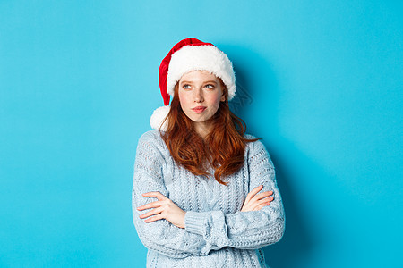 圣诞帽女孩寒假和平安夜的概念 身穿圣诞帽和毛衣的体贴的红发女人 向左看 思考 制定圣诞计划 站在蓝色背景上 时尚 庆典背景