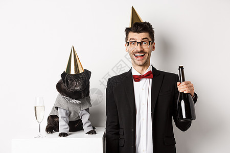 和狗一起欢庆快乐的年轻人 穿着生日晚会花锥子 拿着香槟的人 站在白色背景上 可爱的黑猪和嬉皮士 手 男性图片