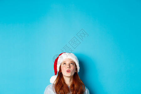圣诞帽女孩冬季假期和圣诞节前夕概念 穿着圣塔帽的漂亮红发女孩的头头从底部出现 抬头看印象深刻的标志 看到宣传品 蓝背景 冬天 迷人的背景