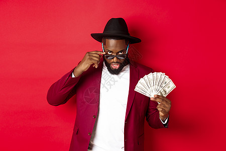 穿着帽子和党服的黑黑男子 展示美元 从太阳眼镜下看 站在红色背景上站立于红底 现金 胡须图片