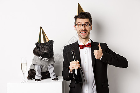 小黑狗戴着党帽 站在欢庆节日的快乐男子旁边 主人举起拇指 拿着香槟瓶子 白背景 乐趣 美丽的图片