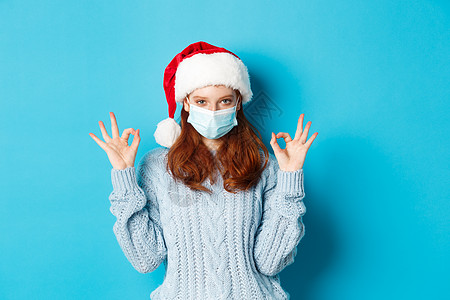 圣诞帽女孩圣诞节 检疫和 covid-19 的概念 戴着圣诞帽和毛衣的可爱红头发少女 戴着冠状病毒的面罩 表现出好的迹象 赞同和赞美某事背景