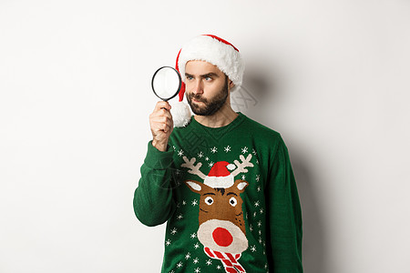 圣诞和节假日概念 穿着圣诞老人帽子的年轻男子在用放大玻璃寻找某种东西 站在白色背景之上 圣诞节 购物图片