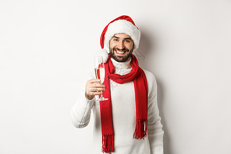 圣诞晚会和节日概念 有胡子的英俊年轻男子 在新年前夕举杯敬酒 拿着香槟和微笑 白背景 冬天 圣诞帽图片
