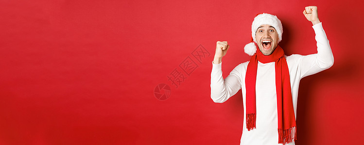欢乐的男子在圣塔帽和围巾中 呼喊欢乐 举起手 庆祝胜利或胜利 战胜红色背景 喜悦 圣诞节图片