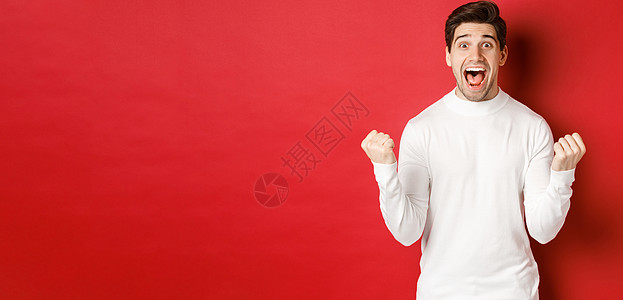 穿着白色毛衣的快乐帅哥的形象 赢得了一些东西 握拳 微笑着惊讶 庆祝胜利 站在红色背景上 冬天 优胜者图片