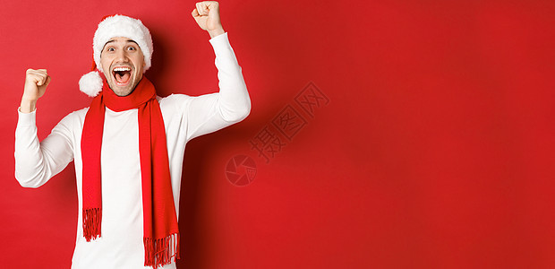 欢乐的男子在圣塔帽和围巾中 呼喊欢乐 举起手 庆祝胜利或胜利 战胜红色背景 工作室 圣诞帽图片